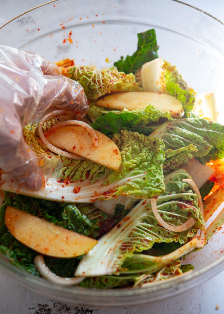 Hand tossing fresh Bomdong cabbage and apple slices with spicy chili dressing in a glass bowl