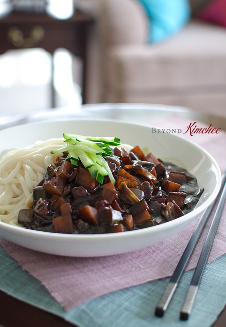 White noodles and black bean jjajangmyeon sauce are garnished with cucumber slices.