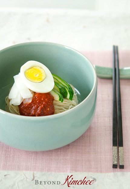 Cold Korean noodles are nesting in a bowl with spicy sauce, pickled radish, cucumber, and hard boiled egg.