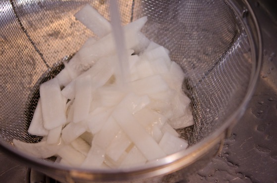 Pickled radish strips are being rinsed in a colander under the running water.