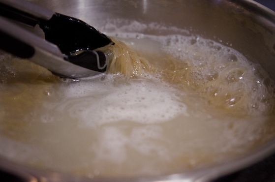 Kitchen tongs are stirring the noodles in boiling water in a pot.