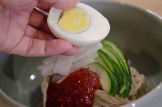 A hard boiled egg sliced in half is added to the top of nangmyeon noodles in a bowl.