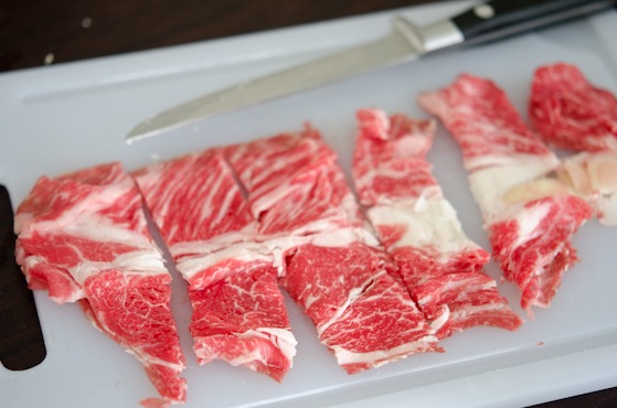 Well marbled beef is thinly sliced on a cutting board.