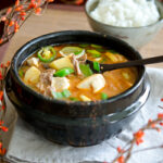 A spoon is dishing up doenajng jjigae with beef and vegetables from the stone pot to show.