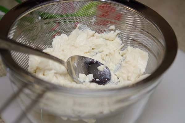 A spoon is smashing the silken tofu through strainer into a mixing bowl.