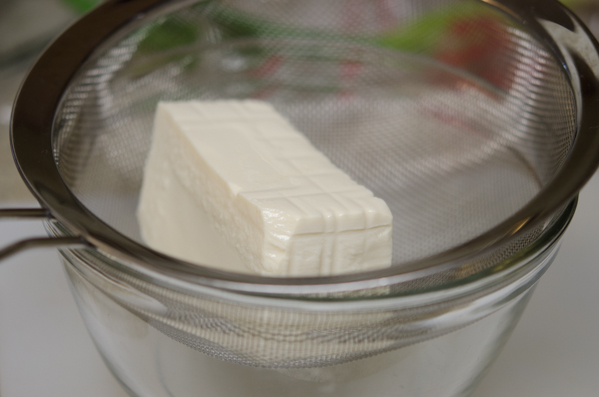 A block of silken tofu is placed on a mesh strainer over a mixing bowl.