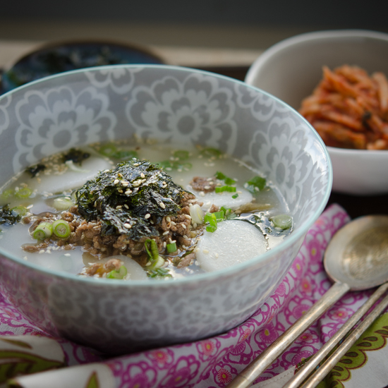 A bowl of rice cake soup is garnished with ground beef sauce and crumbled seaweed.