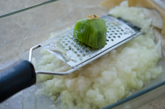 Peeled half kiwi is being grated on a microplane grater.