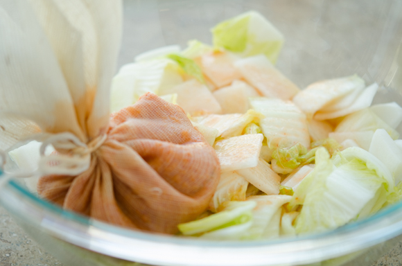 A linen sack of kimchi filling is placed on top of cabbage and radish in a bowl.