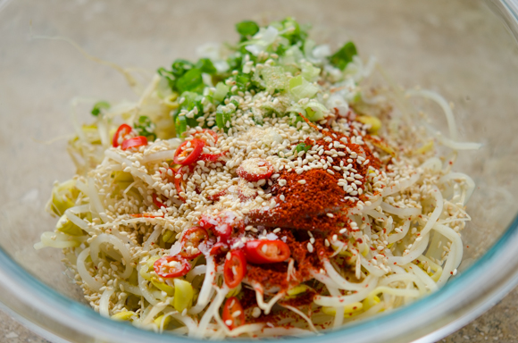 Korean chili flakes and other seasoning ingredients are added to cooked soybean sprouts in a bowl.