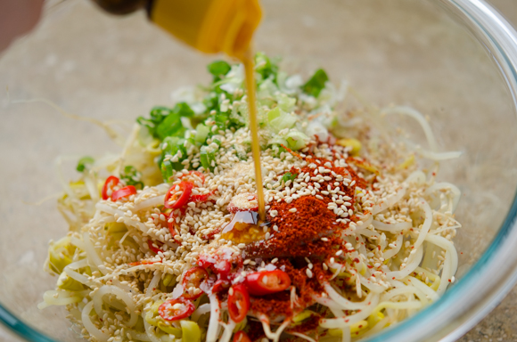 Sesame oil is adding to the soybean sprouts and seasonings in a bowl.