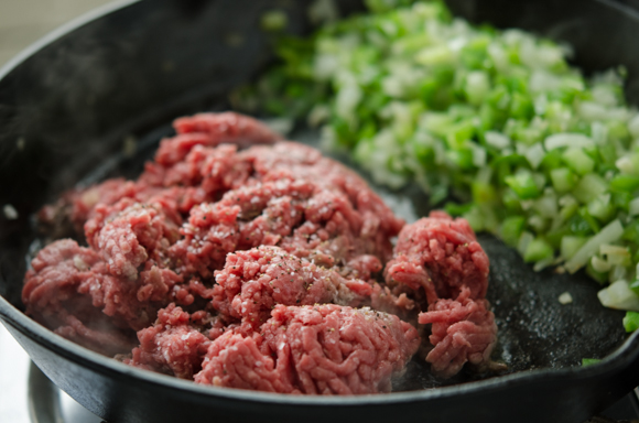 Ground beef is added to the skillet next to vegetables and seasoned with salt and pepper.