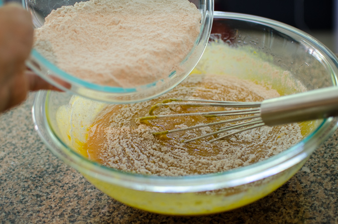 Dry ingredients are added to wet ingredients in a mixing bowl with a whisk.