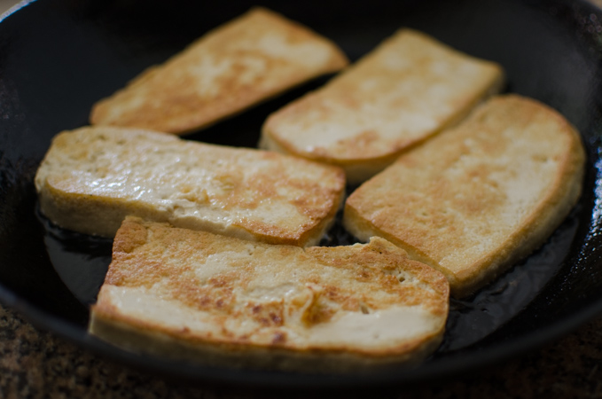 Tofu slices are pan-fried to golden in a skillet.