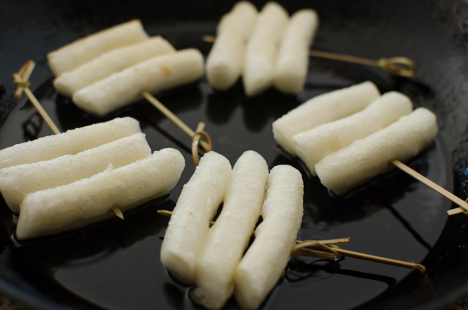 Rice cakes skewered in a bamboo skewers are pan-frying in a skillet.