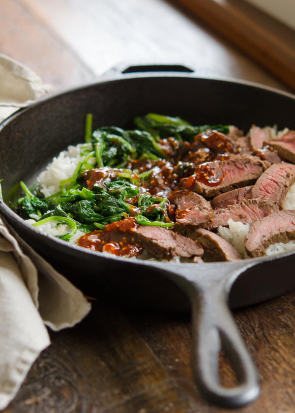 Rice, spinach, beef steak slices are covered with bibimbap sauce in a skillet.