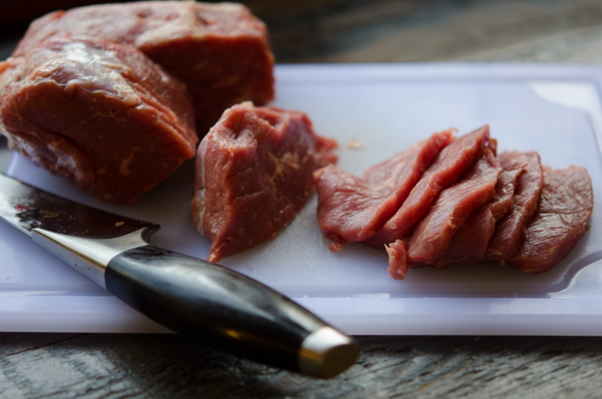 A chunk of lean beef is thinly sliced on a white cutting board.