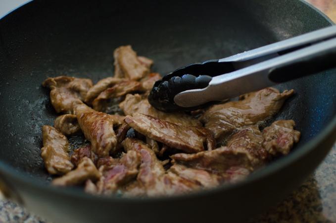 Marinated beef is quickly stir-fried in a wok with kitchen tongs.