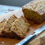 A loaf of carrot oat bread bread is sliced with a knife on a cutting board.