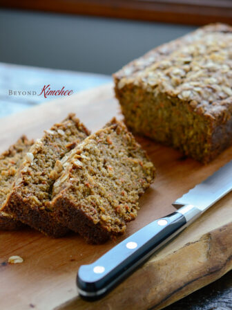 A loaf of carrot oat bread bread is sliced with a knife on a cutting board.