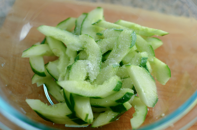 Thinly sliced cucumbers are sprinkled with salt in a bowl.