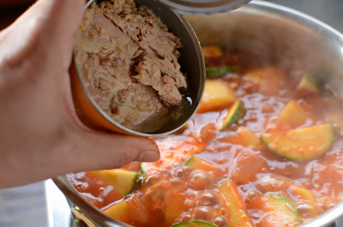 A can of tuna is added to a spicy stew in a pot.