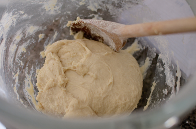 A dough is resting in a glass mixing bowl with a wooden spoon.