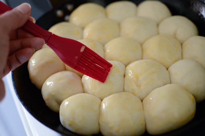 A red brush is applying egg wash to risen beehive shaped buns in a skillet.