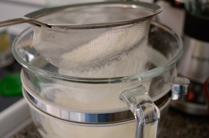 Dry ingredients are being sifted through a mesh strainer over a mixing bowl.