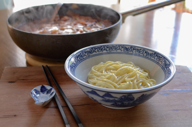 Fresh cooked noodles are placed in a blue bowl in front of a wok filled with jjamppong soup.