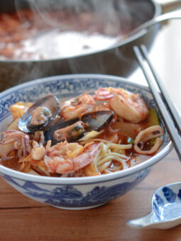 A bowl of noodles are topped with mussel, shrimp, and squid in the spicy soup broth.