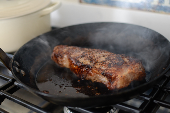 A piece of steak is searing in a skillet on the stove.