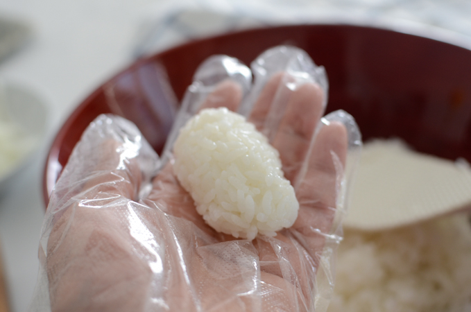 A hand wearing a plastic disposable glove is shaping rice into an oval bite piece.