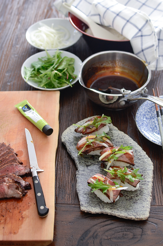 Beef steak slices and a pan of teriyaki sauce is around the adorned beef sushi plated on a stone dish.