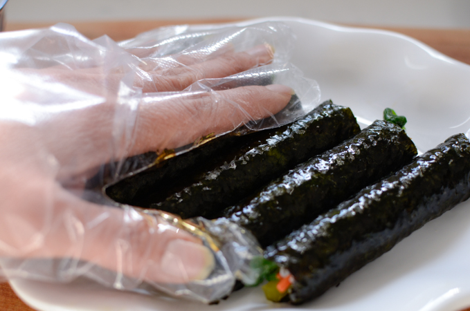 A hand wearing plastic glove is spreading sesame oil on top of mini seaweed rolls.