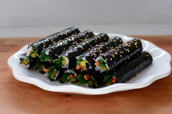 Rows of mini seaweed rolls garnished with sesame seeds are placed on a white serving dish.