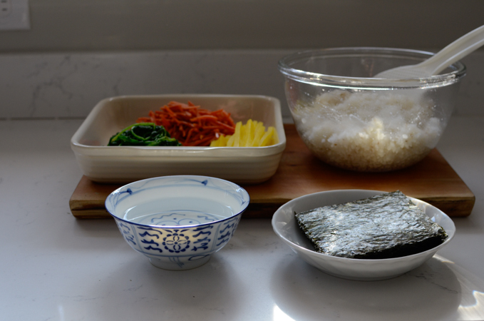 Rice, vegetables, seaweed sheets, and a bowl of water is placed together on the counter.
