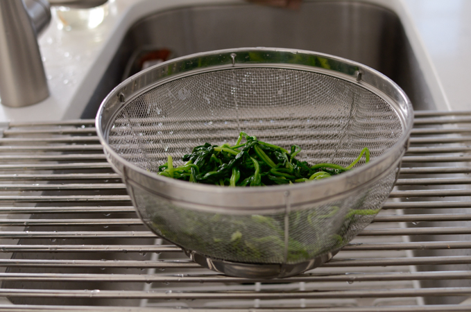 Blanched spinach is drained in a mesh colander and resting on a kitchen rack.