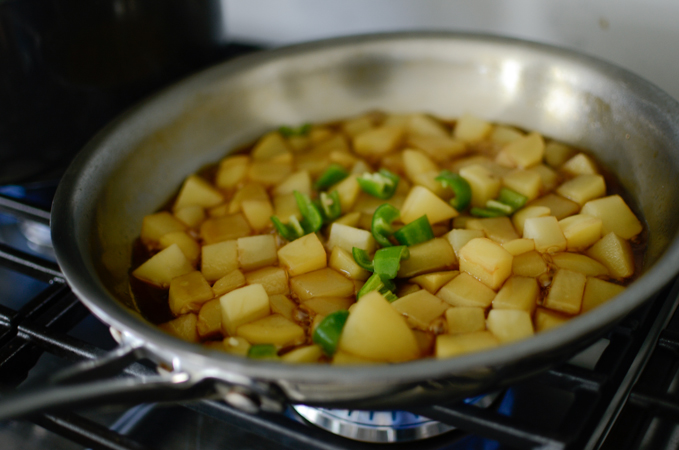 Slices of green chili are added to simmering potato in a skillet.