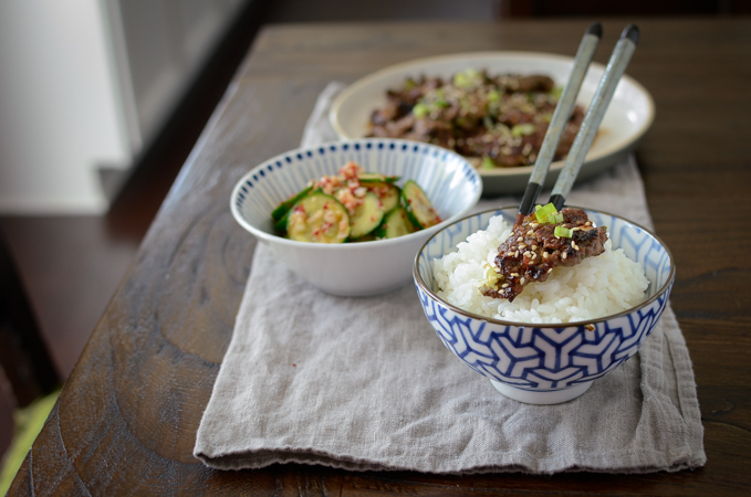 A chunk of crispy bulgogi is topping on a bowl of rice served with cucumber salad on the side.