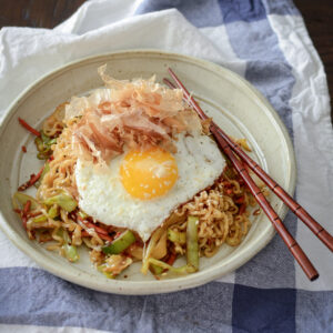 Quick ramen and vegetable stir-fry is topped with fried egg and bonito flakes in a bowl.