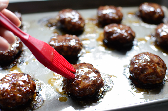 A small red brush is coating the honey glaze onto the cooked beef patties on a baking sheet.