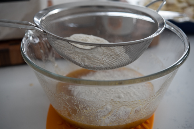 A mesh strainer is sifting flour mixture into the wet mixture in a bowl.