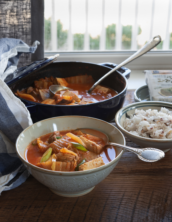 A bowl of kimchi stew made with tomatoes and tofu is served next to rice