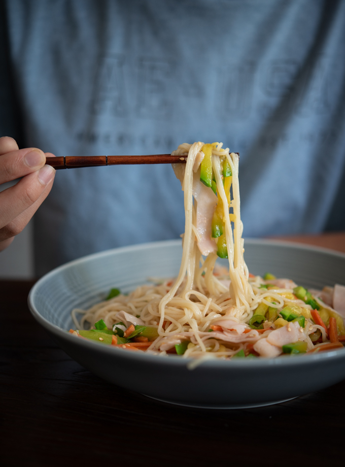 Scooping noodles with deli slices and vegetables with a pair of chopstick.