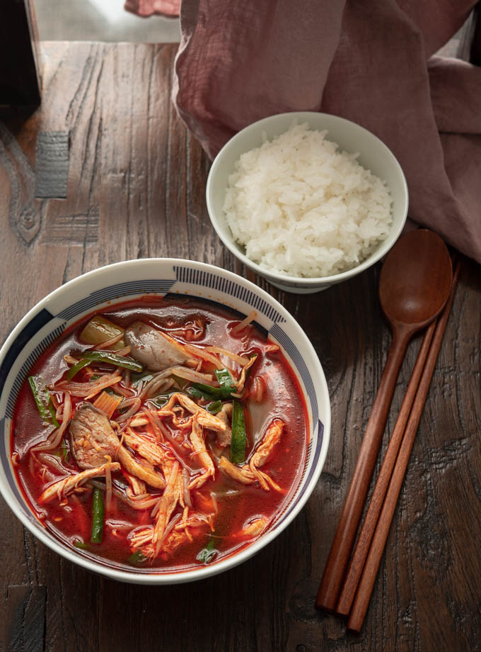 Spicy chicken soup is served in a bowl with rice next to spoon and chopstick.