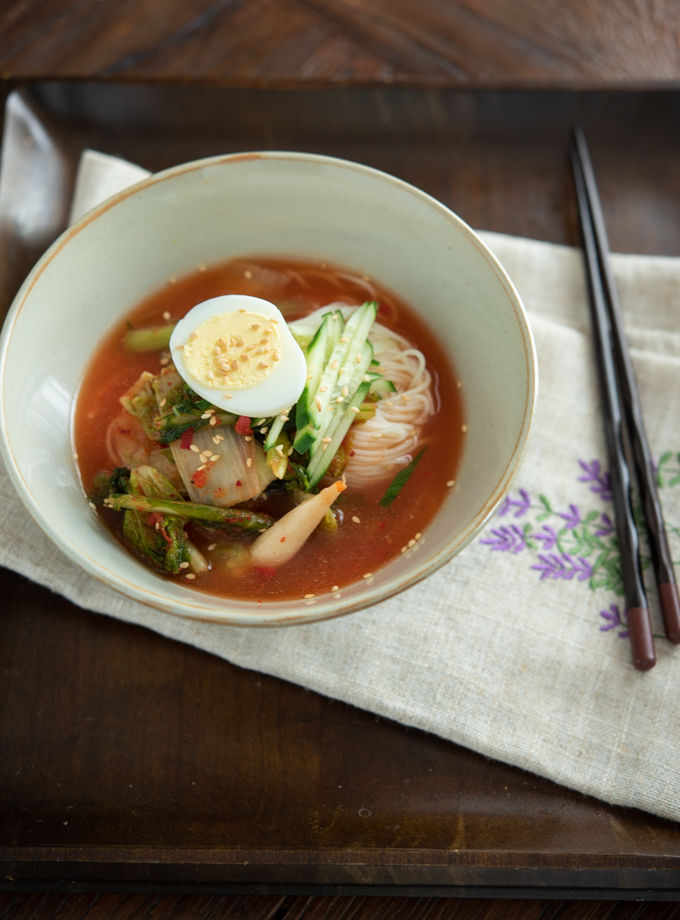 A bowl of noodles are mixed with kimchi and its broth, topped with boiled egg and cucumber.