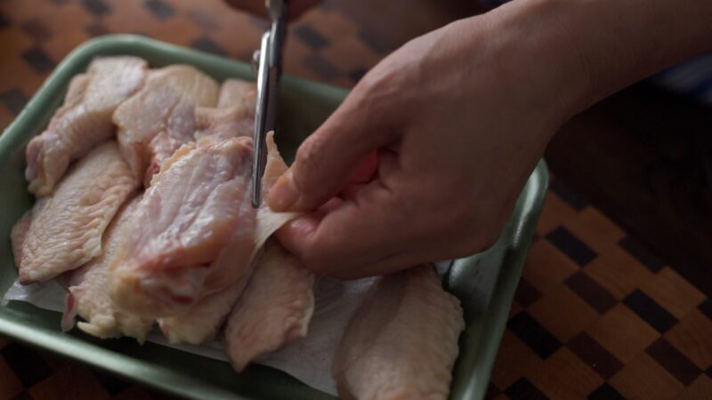 Extra loose skin of chicken party wings are cut off by a pair of scissor.