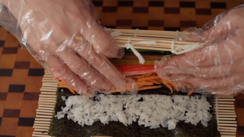 Hands are rolling the bamboo mat to tightly roll kimbap inside.