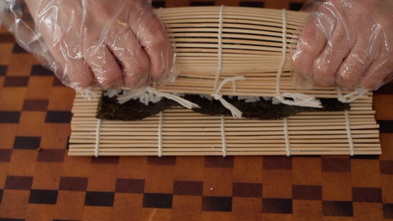 Hands are squeezing the bamboo rolling mat lightly after it is rolled to secure kimbap inside.